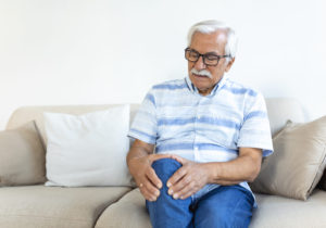 Elderly man sitting on a sofa at home and touching his painful knee. people, health care and problem concept - unhappy senior man suffering from knee ache at home